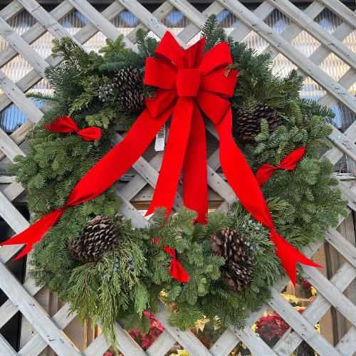 Wreath decorated with a large red ribbon and bow in the center, accented with small red ribbons and pinecones placed around the wreath.