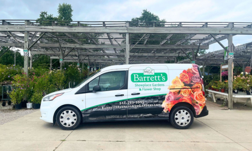 Delivery van with Barrett's Showplace Gardens & Flower Shop branding parked in front of a greenhouse adorned with greenery and flowers. The sky is slightly cloudy but blueish grey. The van displays Barrett's Showplace Gardens & Flower Shop, 517-265-5595, and website link.
